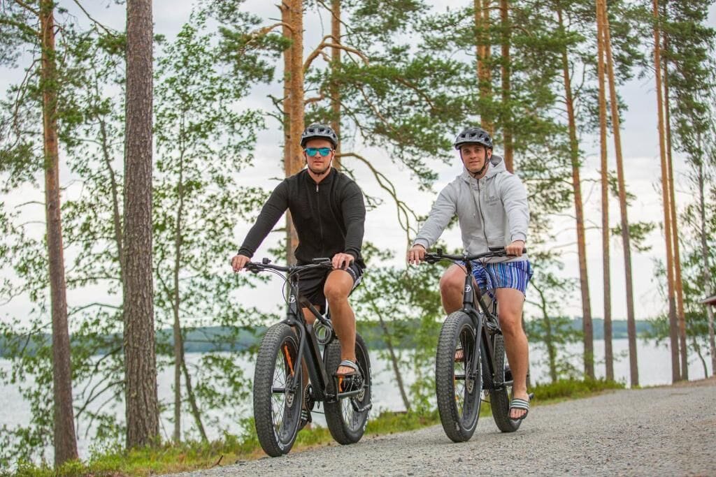 Dos personas en bicicleta en un entorno natural con árboles y un cuerpo de agua al fondo.