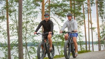 Dos personas en bicicleta en un entorno natural con árboles y un cuerpo de agua al fondo.