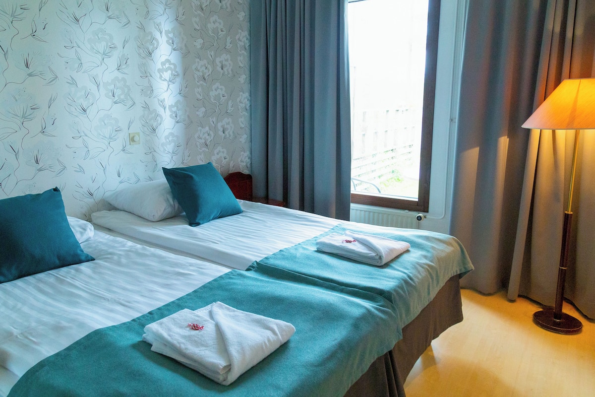 A hotel room with two beds, a window with curtains, a standing lamp, and a floral wallpaper pattern.