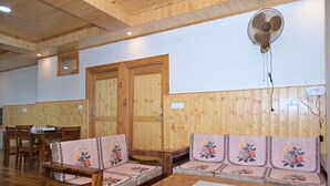 Una habitación con paredes y techo de madera, una mesa de madera, dos sofás de madera con cojines estampados y una puerta de madera, además de un ventilador fijado a la pared.