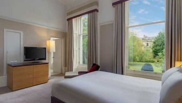 Ein Hotelzimmer mit Bett, Fernseher, Schreibtisch und Fenster mit Blick auf Grünflächen.