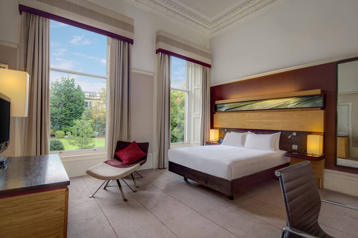 A hotel room with a bed, a desk, a chair, a large window with curtains, and a view of greenery outside.