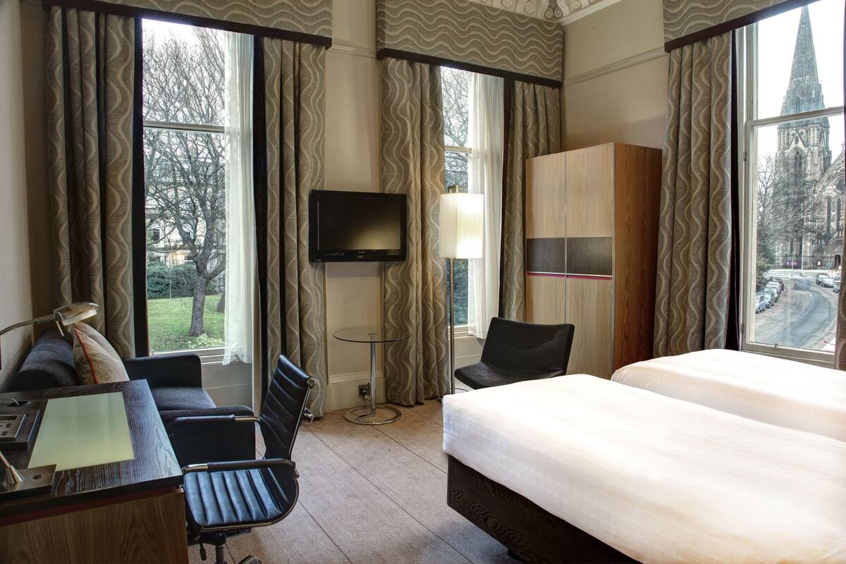 A hotel room with a bed, desk, chair, TV, and large windows with curtains.