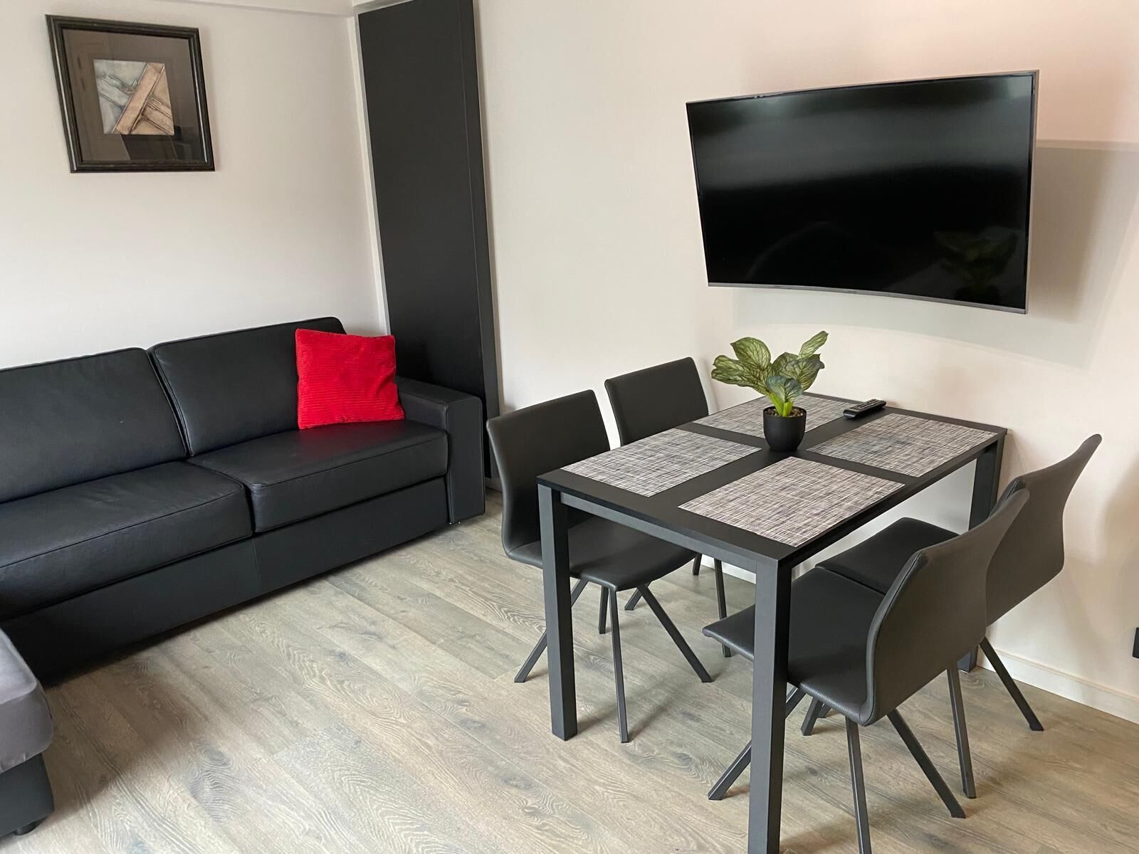 Una sala moderna con un sofá negro, una almohada roja, una mesa de comedor con cuatro sillas, una planta en maceta y un televisor de pantalla plana montado en la pared.