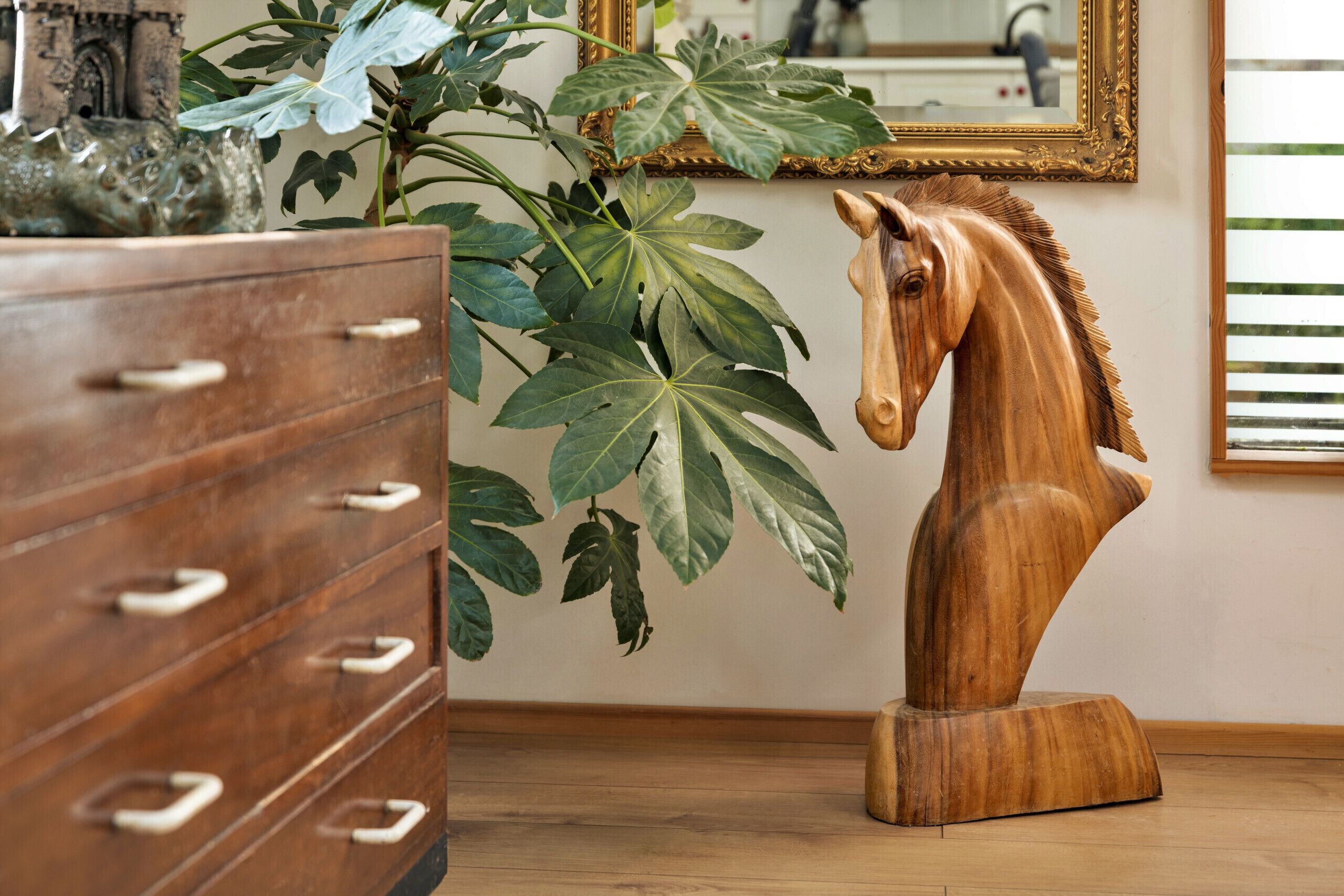 Une commode en bois avec des tiroirs, une grande plante ficus et une sculpture équestre en bois.