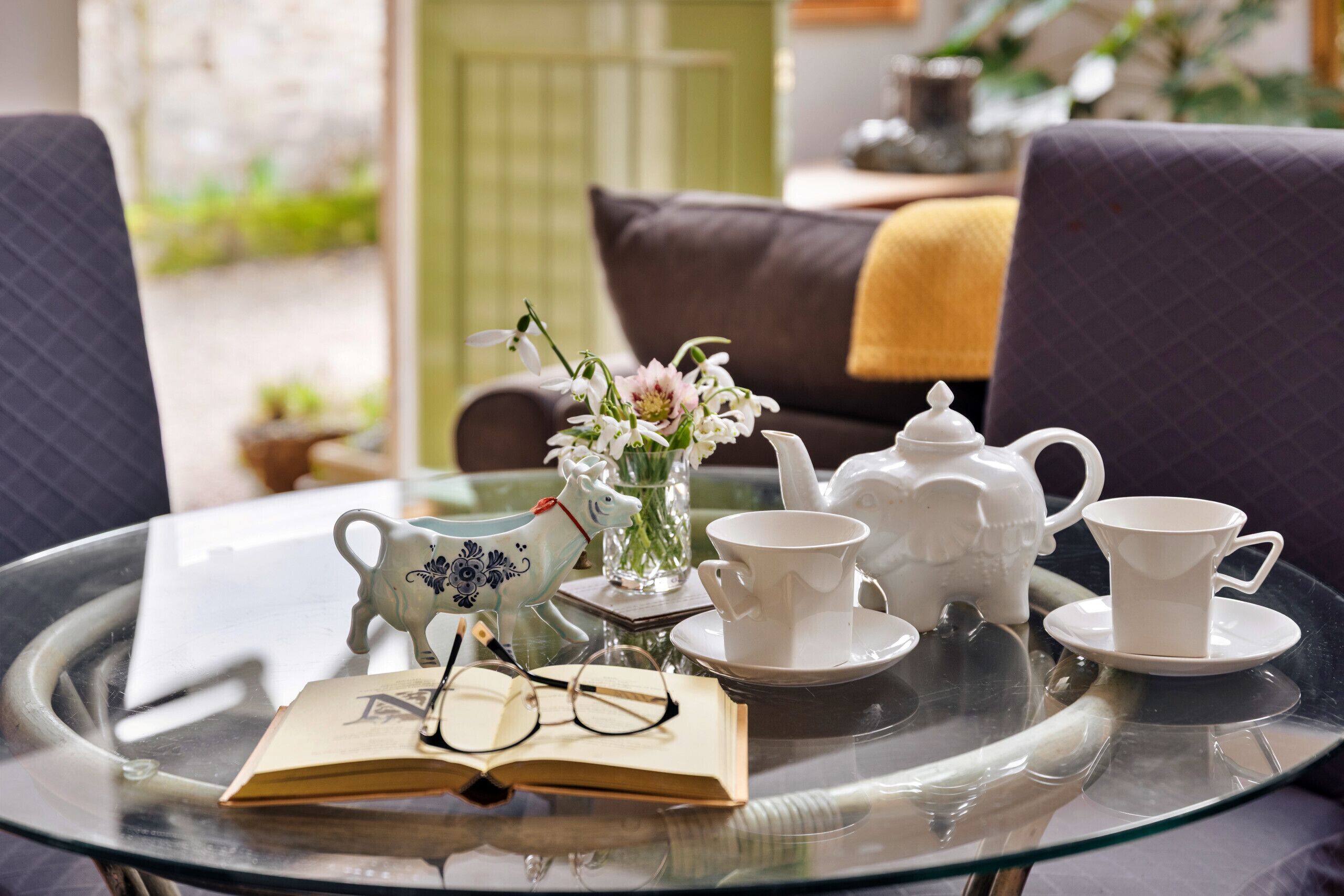 A glass table with a book, teapot, cups, and a decorative horse figurine.