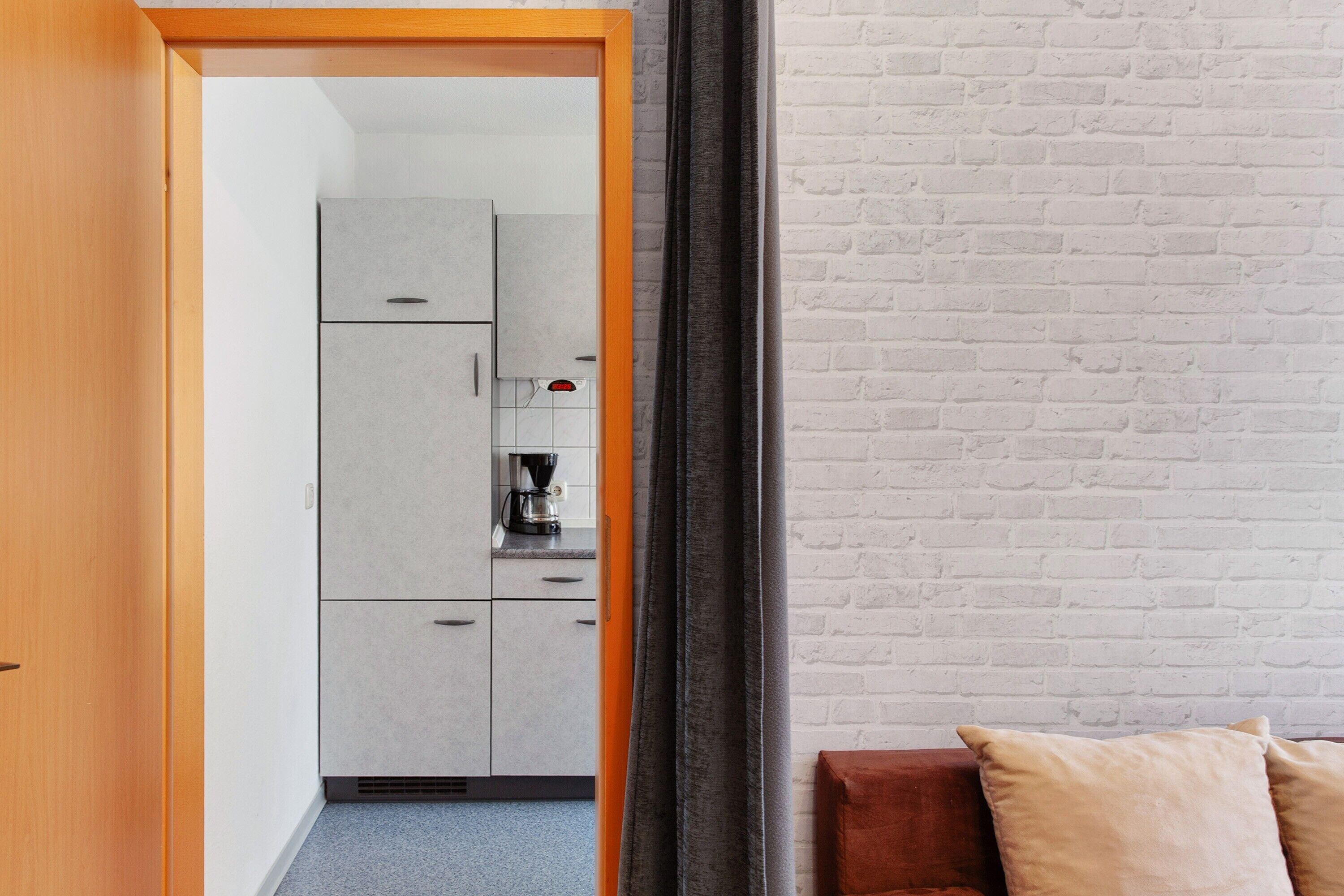 Een kamer met een houten deur, een kitchenette met kastjes en een koffiezetapparaat, en een bakstenen muur.