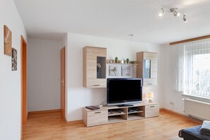 Uma sala de estar moderna com televisão de tela plana, armários de madeira e uma janela com cortinas leves.
