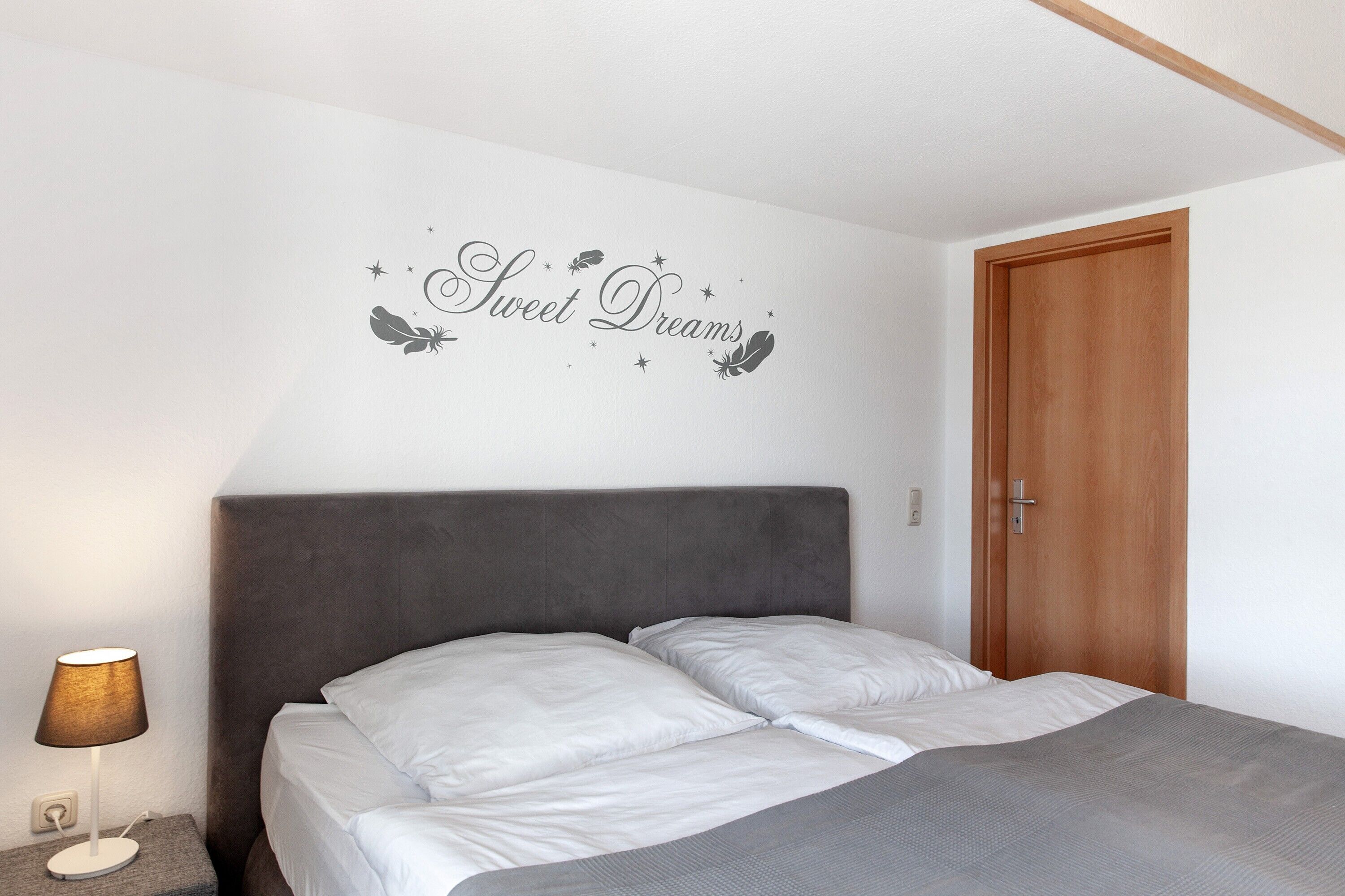 A bedroom with a bed, a nightstand with a lamp, a wooden door, and a wall with the text 'Sweet Dreams'.