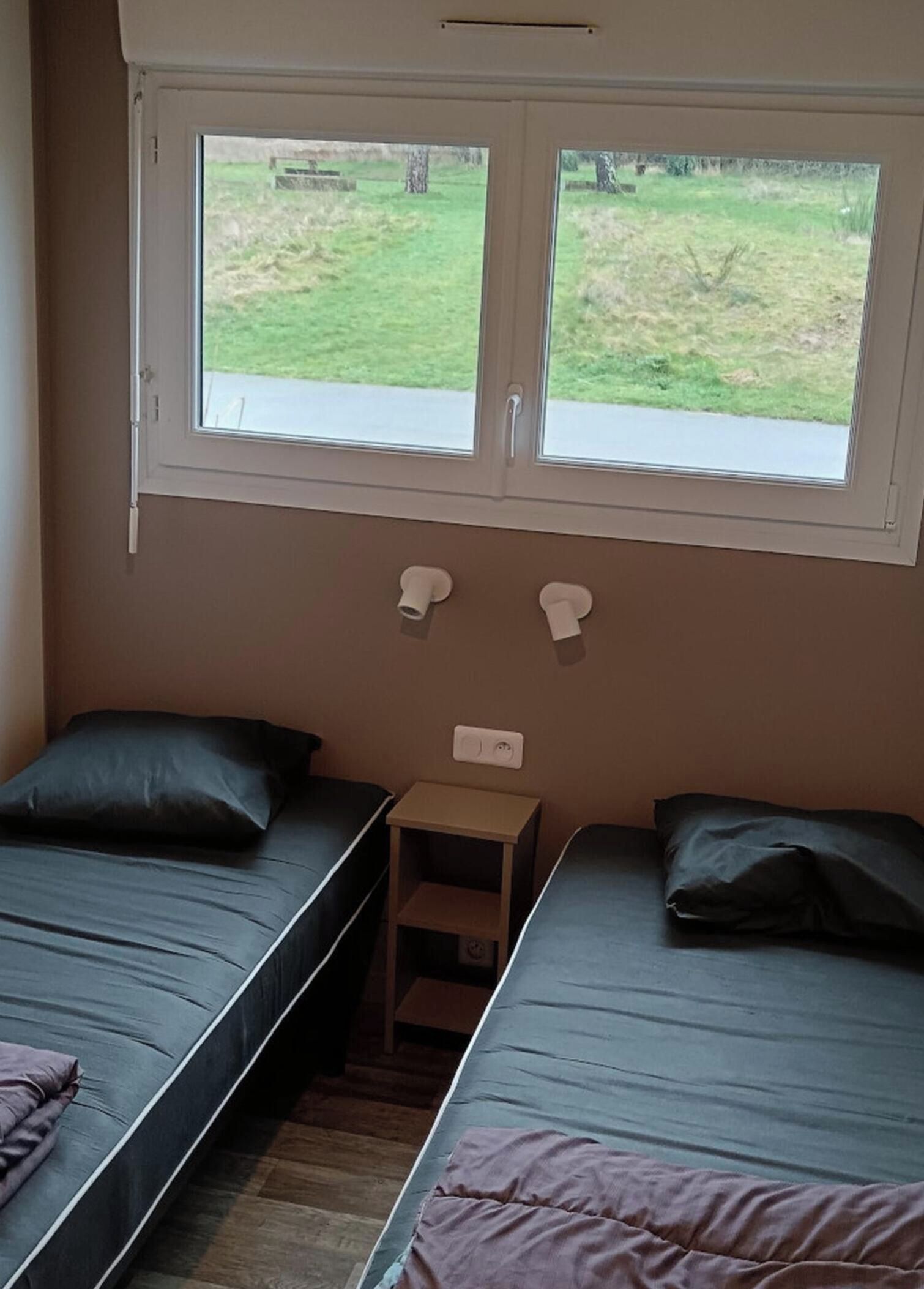 A room with two single beds, a small wooden nightstand, and a window with a view of the outdoors.
