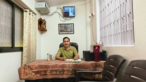 A man sitting at a desk with a computer, papers, and a red water bottle. There is a window with blinds, a wall-mounted air conditioner, and a framed picture on the wall. - Shri Jee Kunj Homestay (Varanasi)