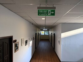 A hallway with a fire exit sign, framed pictures on the walls, and doors leading to rooms.