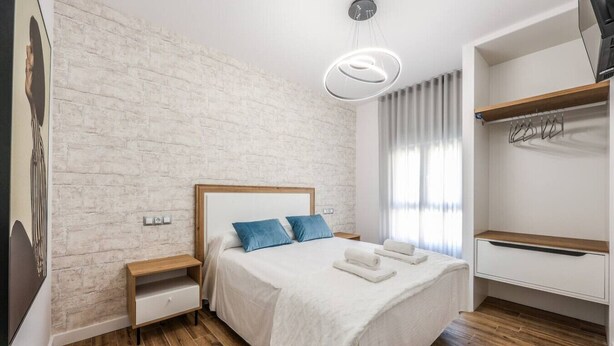 A bedroom with a bed, wooden nightstands, a white brick wall, a hanging light fixture, and a closet with a wooden shelf.