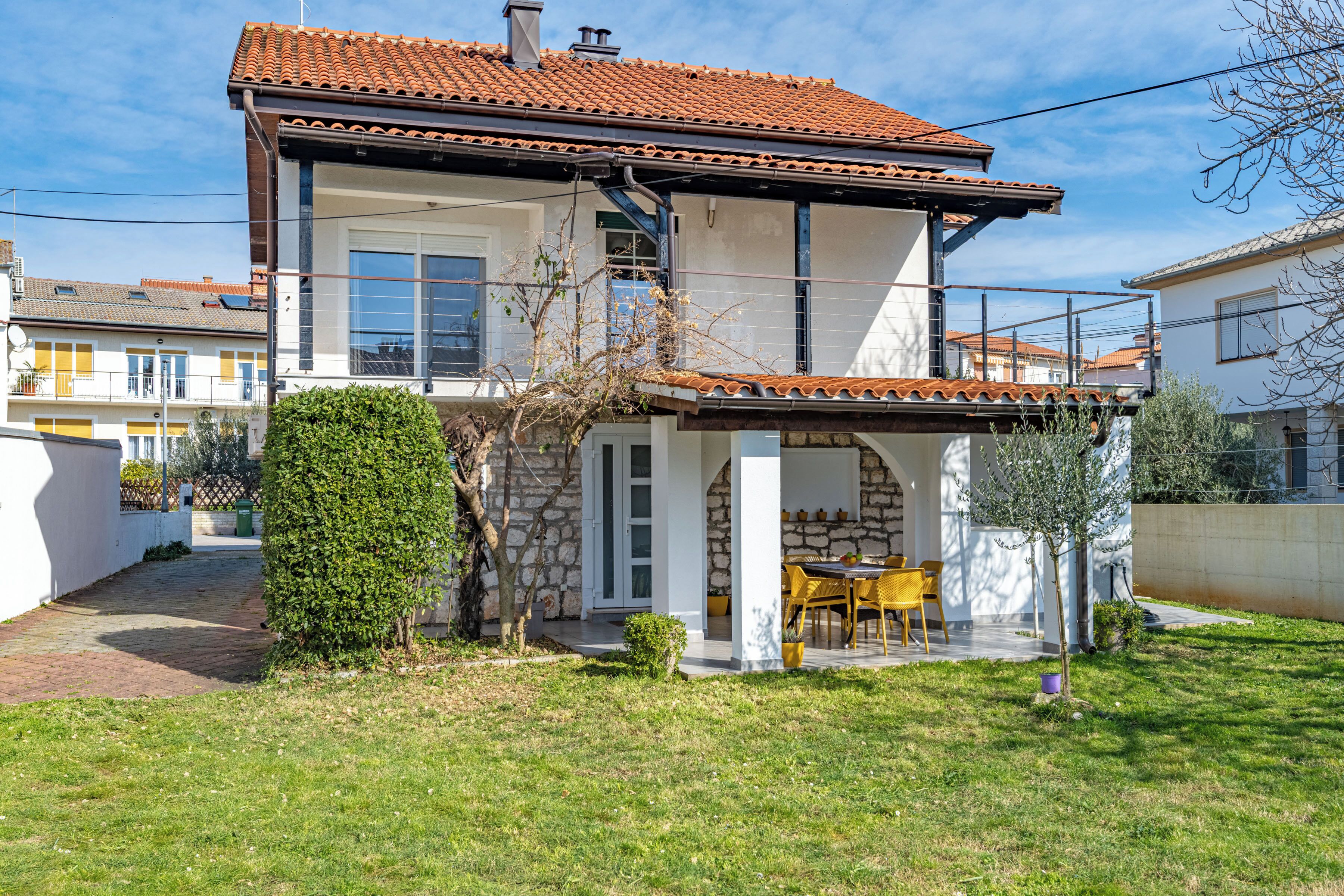 A two-story house with a tiled roof, a covered patio with a table and chairs, and a well-maintained lawn.
