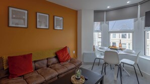 A modern living room with a brown leather sofa, a dining table with chairs, and large windows with a view of buildings. - Bright 3BR Duplex Steps from Shops & Dining (Cardiff)