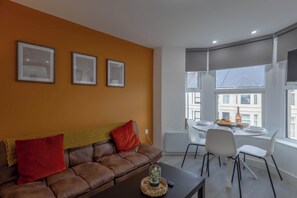 A modern living room with a brown leather sofa, a dining table with chairs, and large windows with a view of buildings. - Bright 3BR Duplex Steps from Shops & Dining (Cardiff)