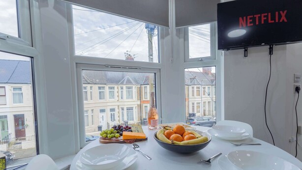 A dining table with plates, cutlery, and a bowl of fruit and cheese. A bottle of wine is on the table. A TV mounted on the wall displays 'NETFLIX'. - Bright 3BR Duplex Steps from Shops & Dining (Cardiff)