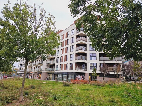 Un immeuble résidentiel à plusieurs étages, doté de balcons, avec un espace verdoyant devant et un arbre sur le côté gauche.