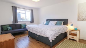A bedroom with a bed, a sofa, a bedside table, a lamp, and a window with curtains. - Snowdrop Cottage (Maidstone)