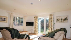A spacious living room with two beige sofas, a flat-screen TV, a small table, and a sliding glass door leading to an outdoor area. - Snowdrop Cottage (Maidstone)