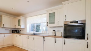 A modern kitchen with white cabinets, a wooden countertop, a built-in oven, and a window with a view of greenery outside. - Snowdrop Cottage (Maidstone)