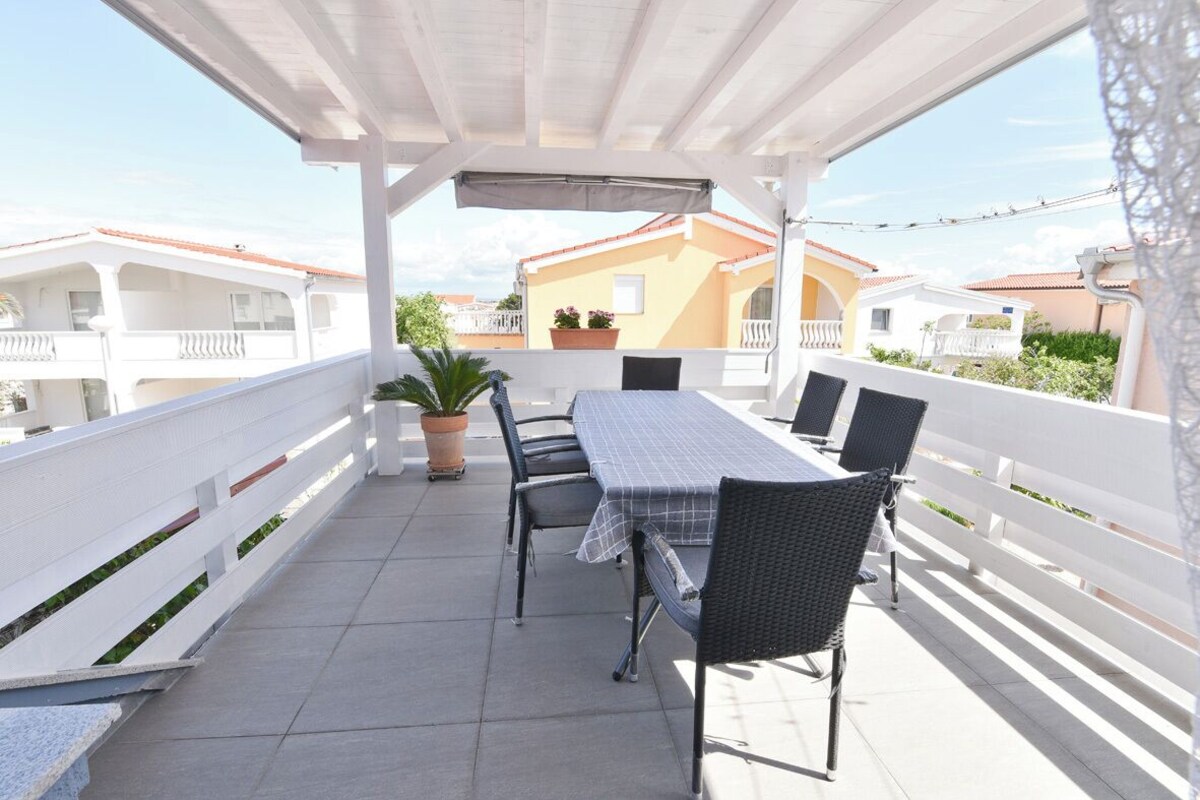 Eine überdachte Terrasse im Freien mit Tisch und Stühlen, Blumentöpfen und Blick auf angrenzende Gebäude.