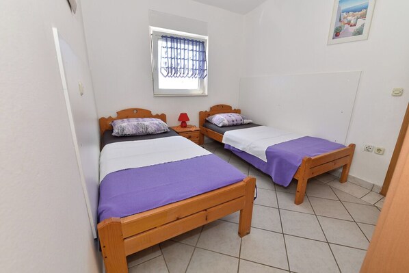 A room with two wooden beds, each with a purple bedspread, a window with blue curtains, and a small red table. - Apartments Karacic (Vir)