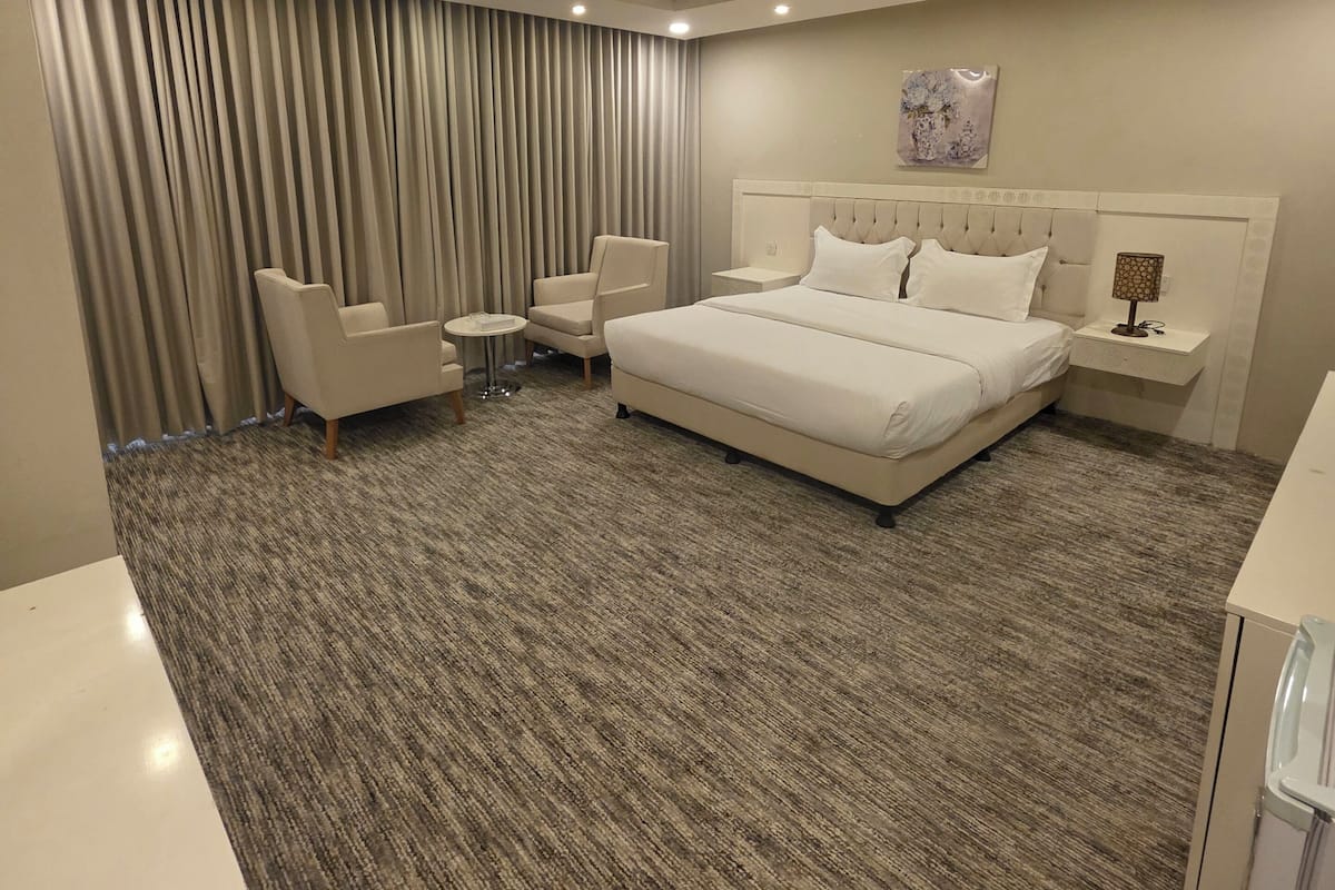 A modern hotel room with a large bed, two armchairs, a small table, and a large window with curtains.