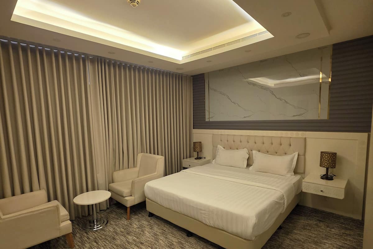 A modern hotel room with a large bed, a seating area with a chair and a small table, a nightstand with a lamp, and a ceiling with recessed lighting.