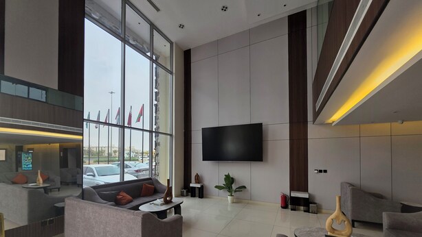 A modern hotel lobby with a large TV, comfortable seating, a reception desk, and a view of parked cars outside. - Palette Ninty (Riyadh)