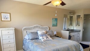 Ein Schlafzimmer mit Bett, Kommode, Deckenventilator und Bad mit Dusche. An der Wand hängt ein gerahmtes Bild.