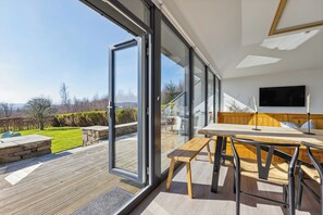 Un intérieur moderne avec une grande porte-fenêtre coulissante donnant sur une terrasse, une table à manger en bois et un téléviseur fixé au mur.