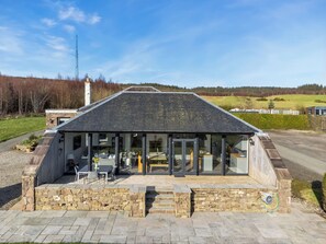 Une maison moderne avec des fondations en pierre, un toit en pente et un patio couvert aménagé avec du mobilier d’extérieur.