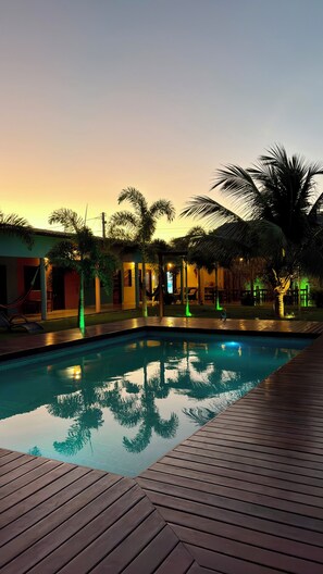 A swimming pool with a wooden deck, surrounded by palm trees and a building with a thatched roof. - The Jungle Guriú (Camocim)