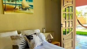 A hotel room with a bed, pillows, a painting of a beach scene, a wooden door, and a view of a garden. - The Jungle Guriú (Camocim)