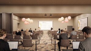 Una sala conferenze con schermo per presentazioni, partecipanti seduti a tavoli con laptop e pavimento rivestito di moquette.