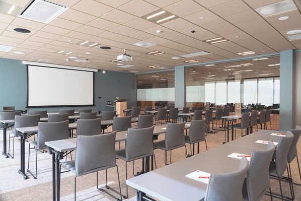 Un salón de conferencias con filas de mesas y sillas, una pantalla de proyector y un ventanal grande.