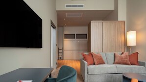 Una sala de estar moderna con un sofá gris, un sillón verde azulado y una pequeña mesa con un bolígrafo rojo. Un televisor de pantalla plana está montado en la pared, y una lámpara se encuentra sobre una mesita de noche.