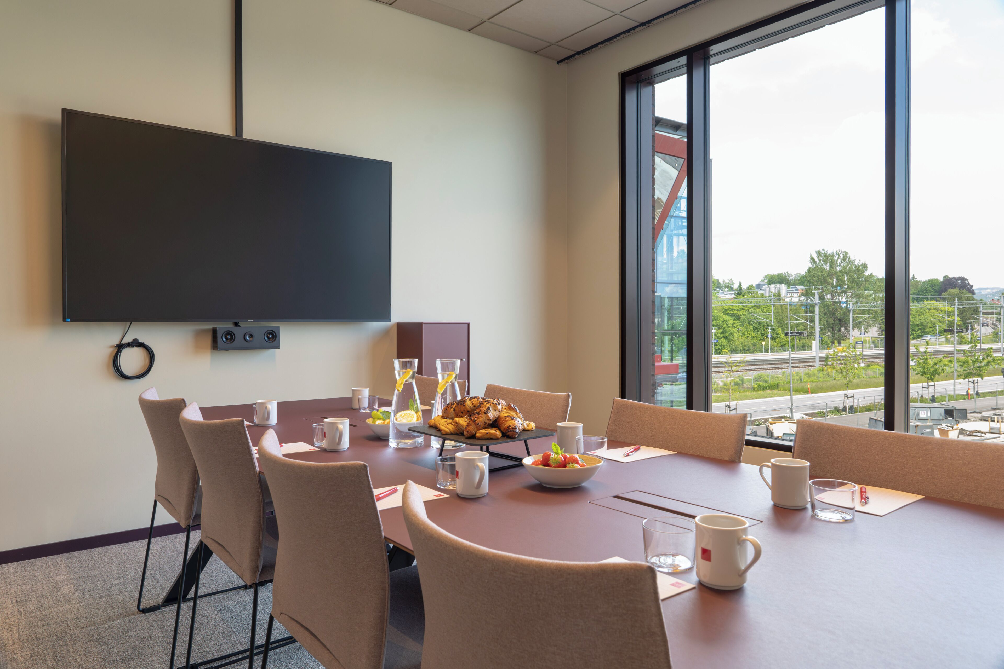 Een vergaderruimte met een grote tafel, stoelen, een flatscreentelevisie en een uitzicht op een stadsgezicht door grote ramen.