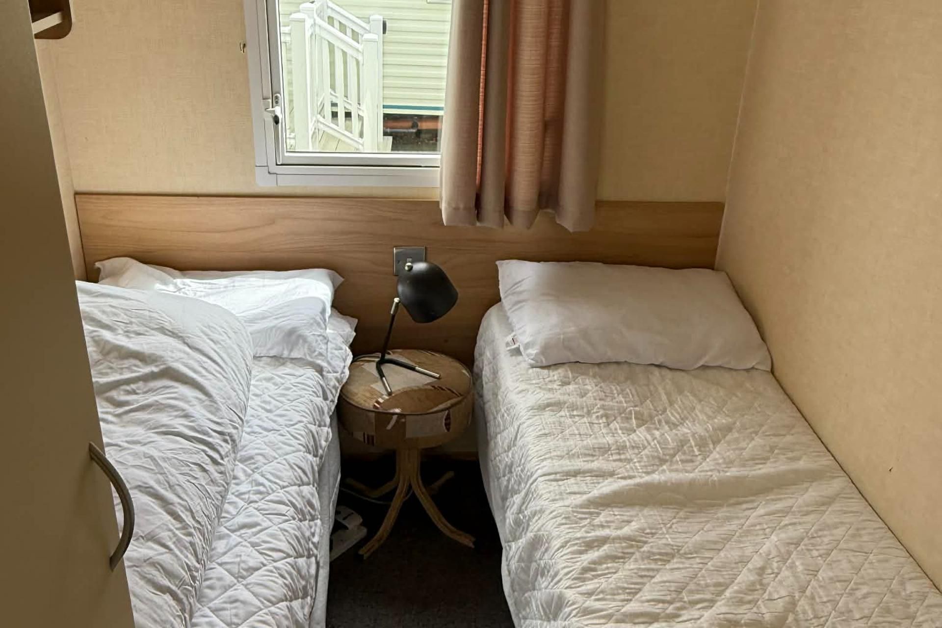 Habitació petita i senzilla amb dos llits individuals, una taula rodona amb una làmpada i una finestra amb cortines.