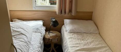Habitació petita i senzilla amb dos llits individuals, una taula rodona amb una làmpada i una finestra amb cortines.
