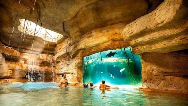 Una piscina interior con una amplia pared de vidrio, paredes rocosas y una cascada.