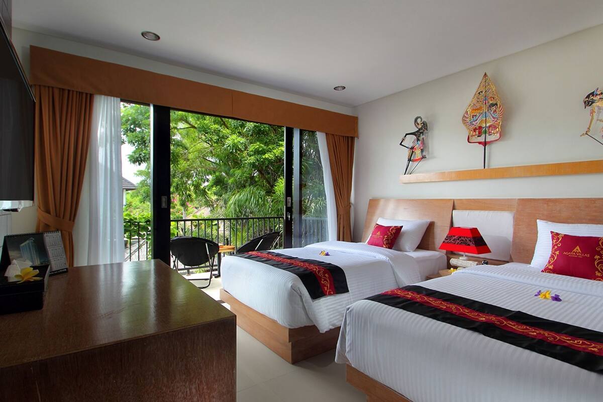 A hotel room with two beds, a desk, a chair, and a balcony with a view of greenery.