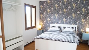 A bedroom with a checkered bedspread, a white headboard, a nightstand with a lamp, and a floral wallpaper. - Casa Florian (Vito d'Asio)