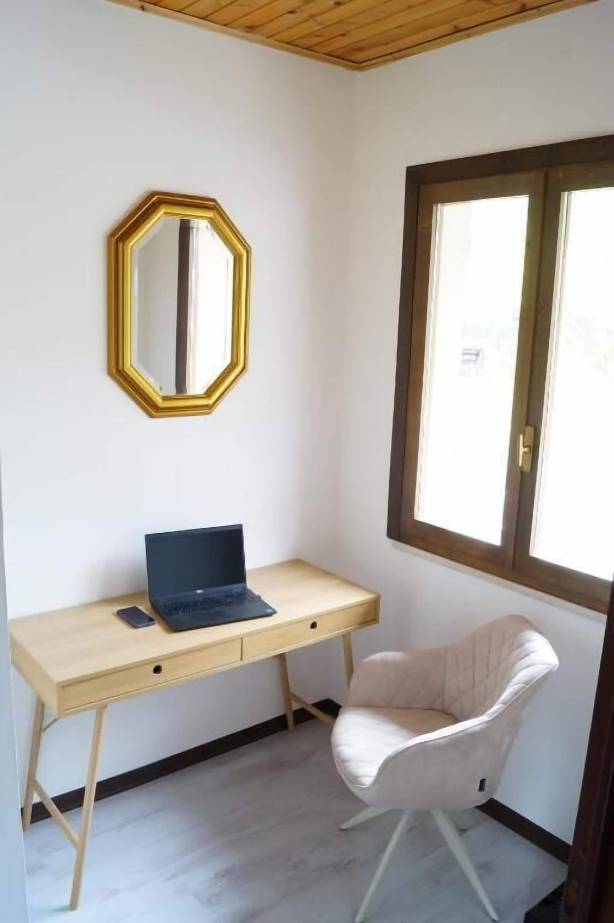 A room with a wooden desk, a laptop, a chair, a mirror, and a window.