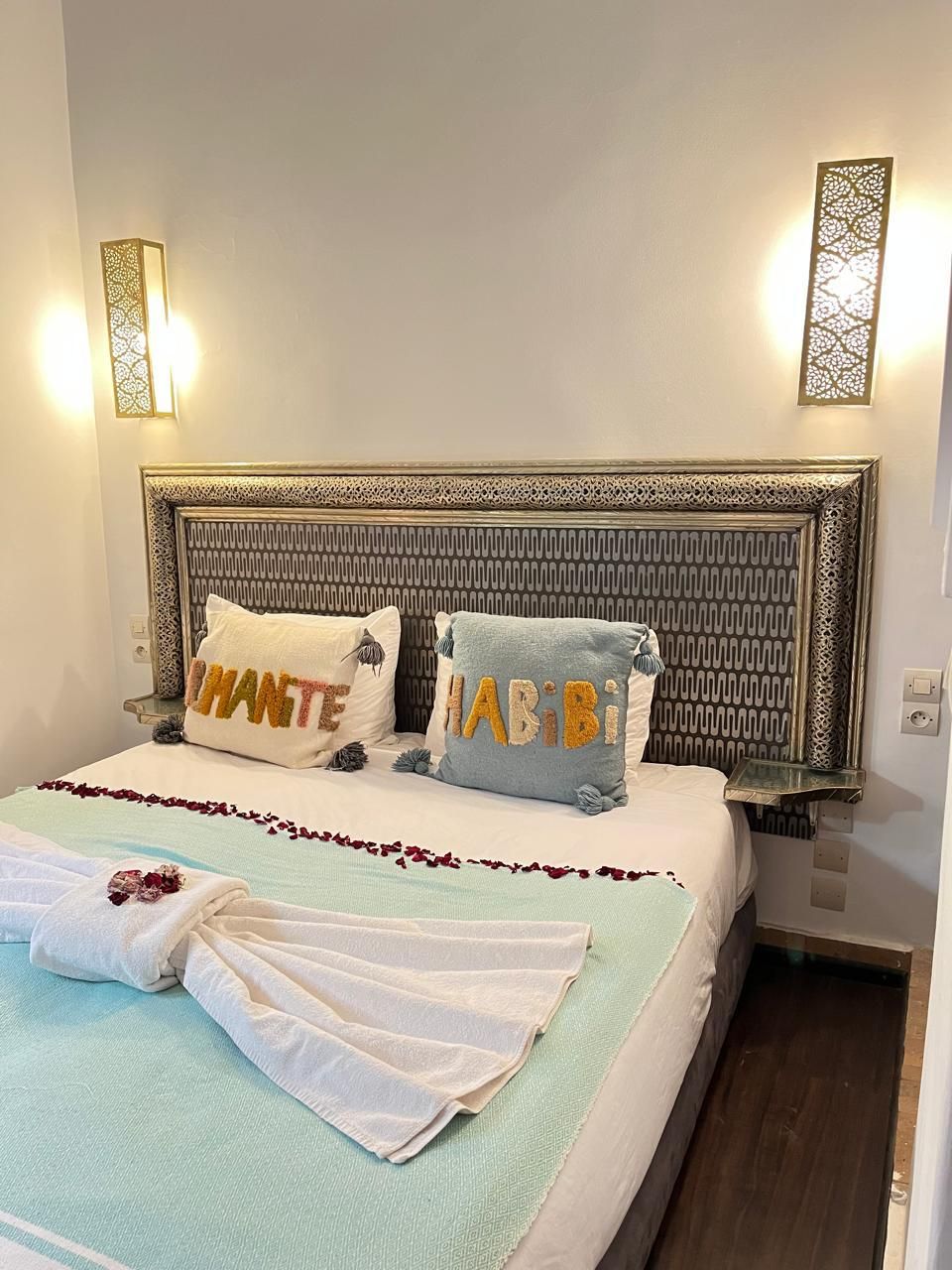 A hotel room with an ornate headboard, two pillows with text, a neatly made bed with a towel, and wall-mounted lights.