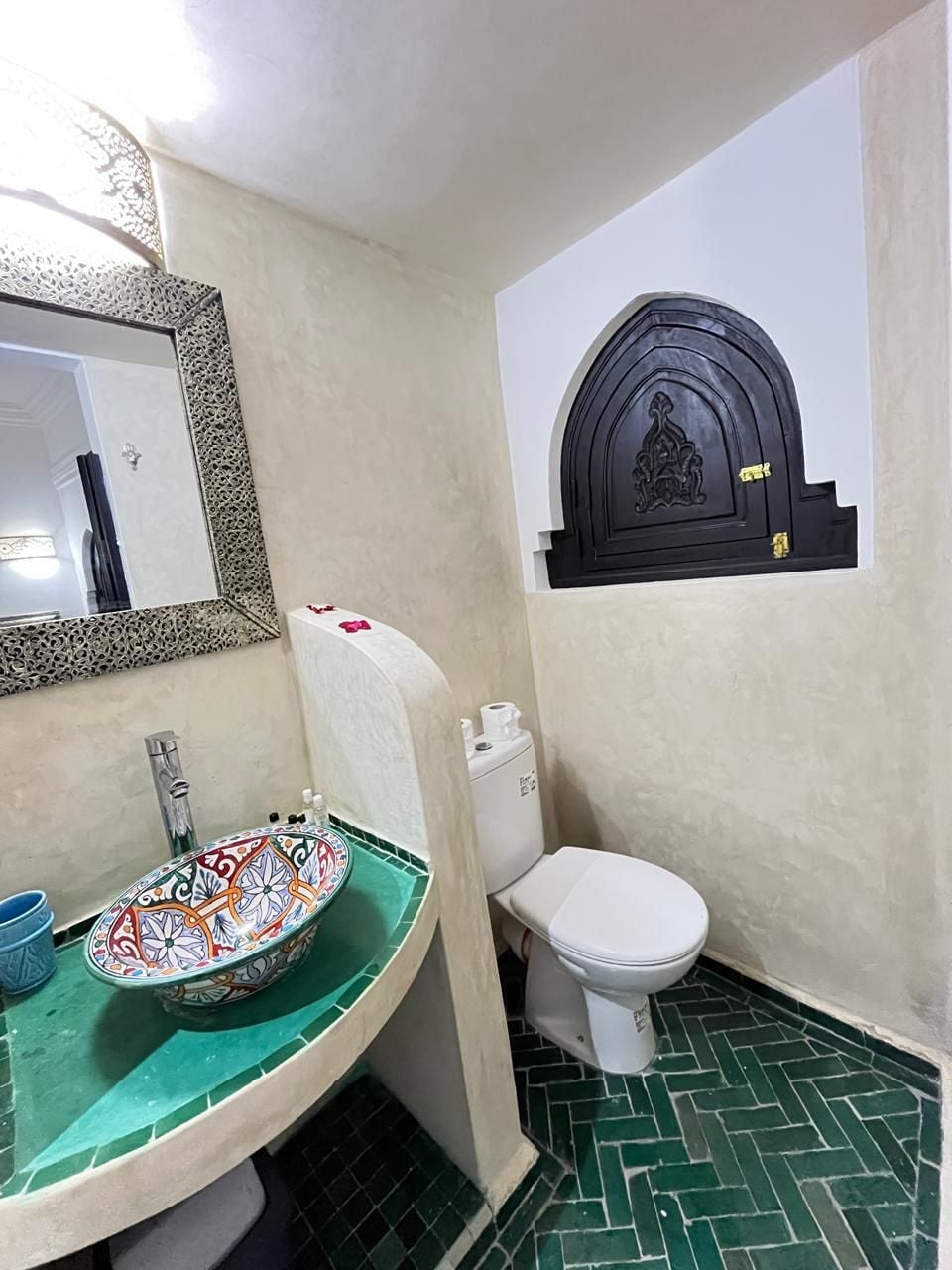 A bathroom with a green tiled floor, a white toilet, a sink with a colorful bowl, and a decorative wall piece above the toilet.