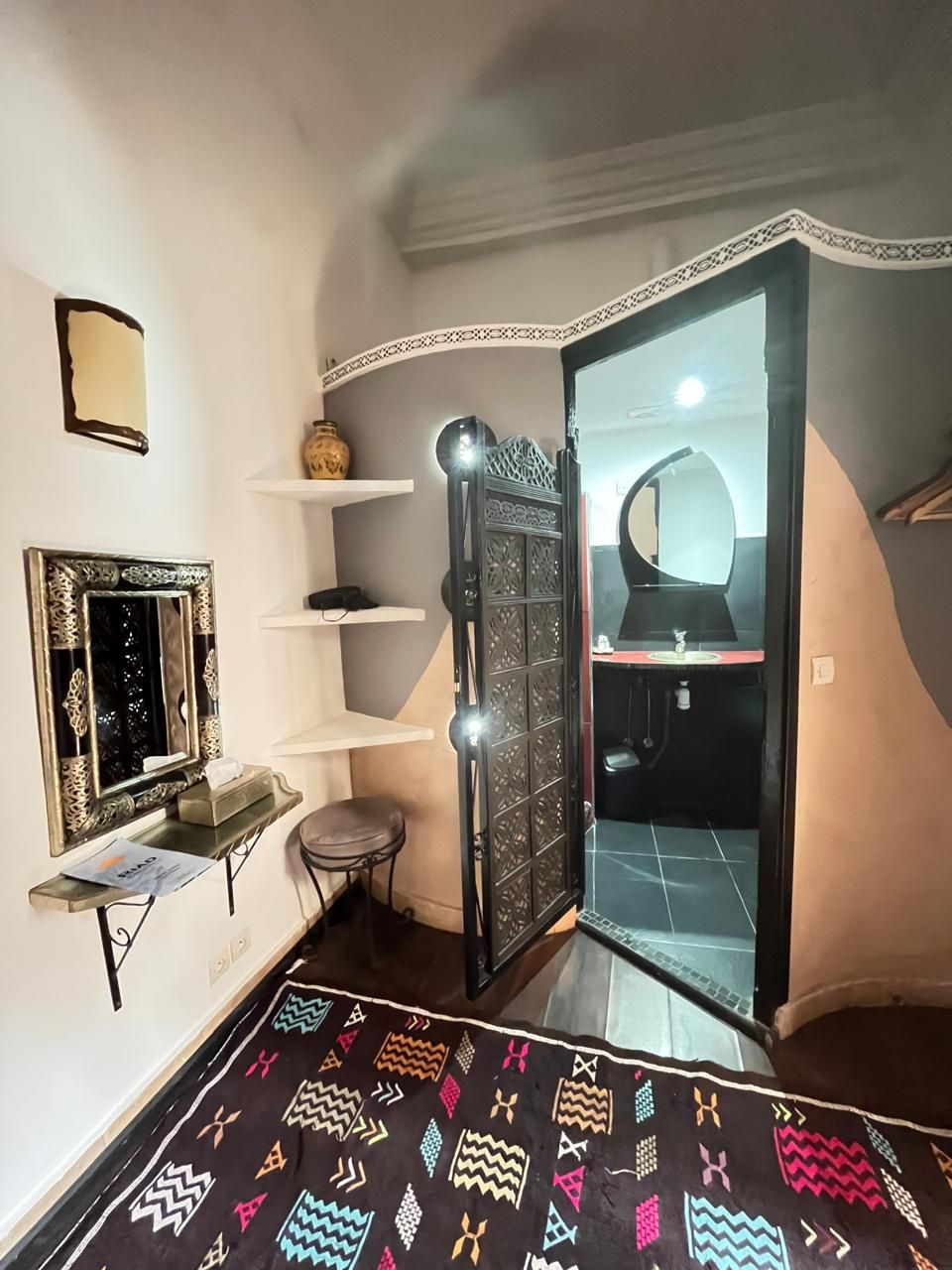 A room with a patterned rug, a mirror, shelves, a cabinet, and a bathroom with a sink and mirror.