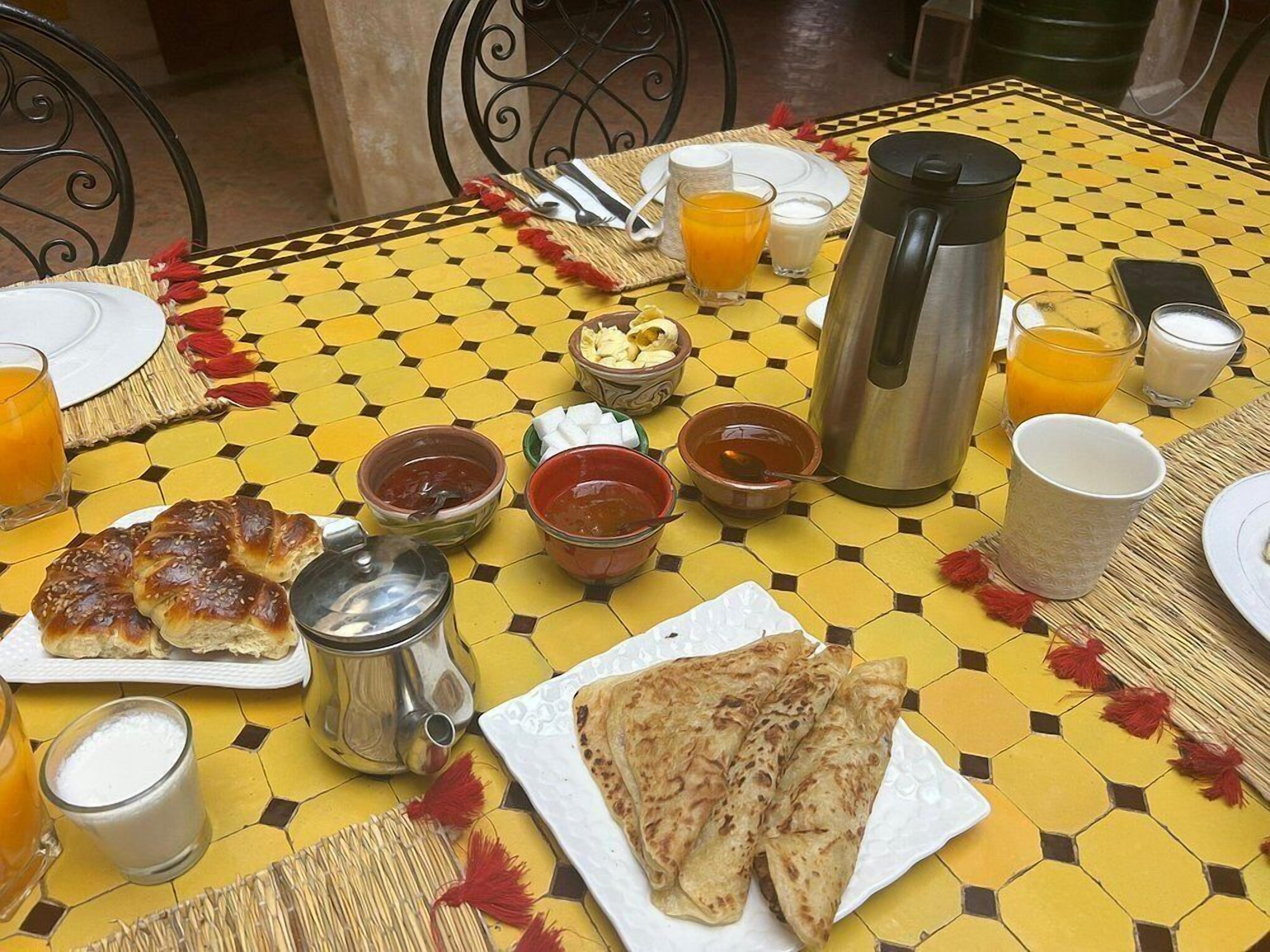 A breakfast table with a yellow patterned tablecloth, a plate of flatbreads, a thermos, and various condiments.