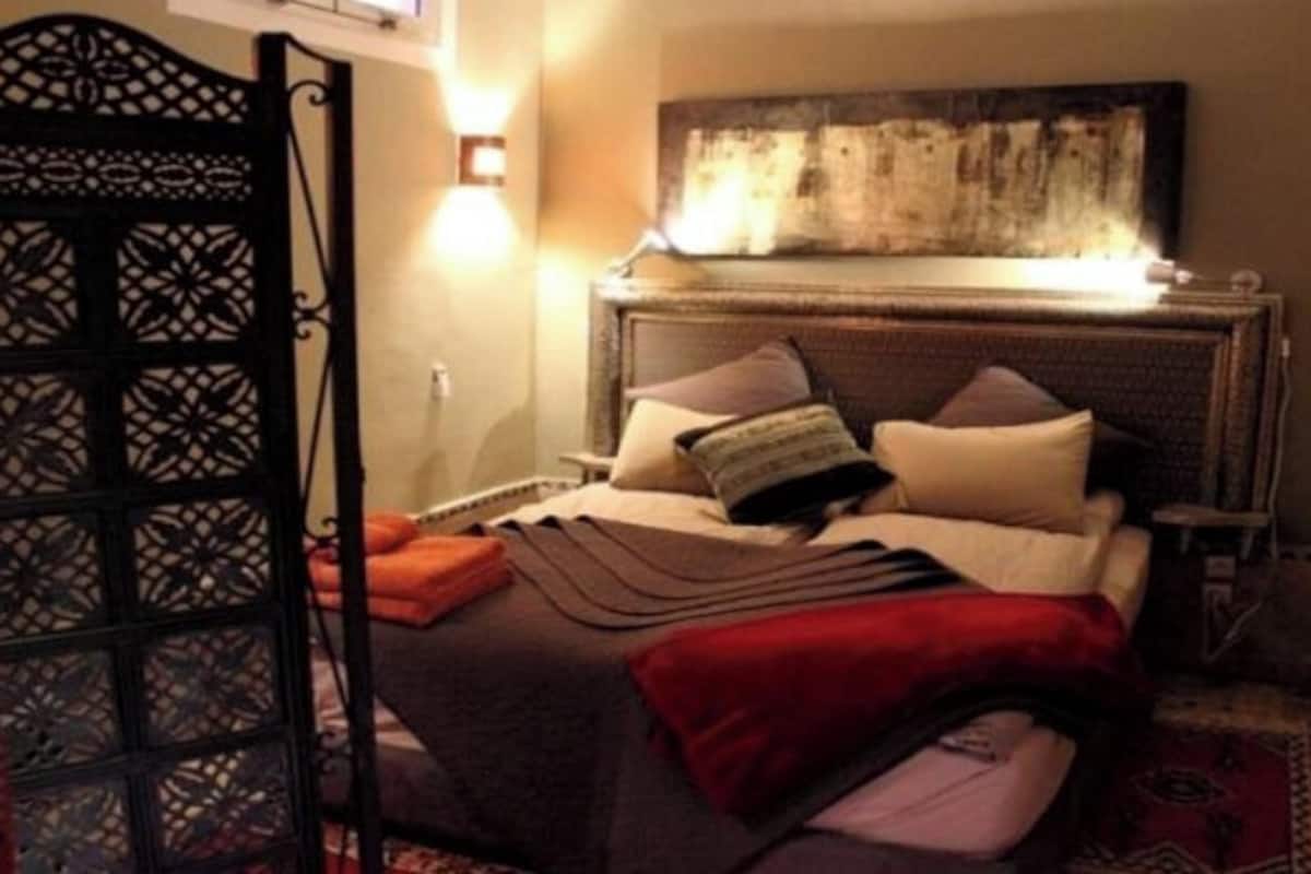 A bedroom with a bed, pillows, a blanket, a wall-mounted artwork, and a door with a decorative pattern.